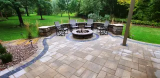 Large paver patio with a curved seat wall, round fire pit, and lounge chairs overlooking a tree-lined lawn